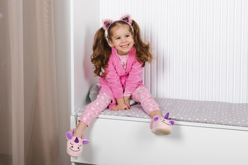 A charming little girl in a pink pajama and robe is sitting on a childrens bed and happily smiling.
