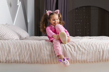 A charming little girl in a pink pajama and robe is sitting on the bed, putting on her soft slippers and smiling.