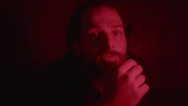Close-Up Shot of a Young, Bearded, Long-Haired Man Looking into the Camera under a Red Light