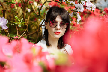 A brunette in a beige dress and glasses among blooming azaleas