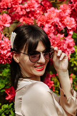 A brunette in a beige dress and glasses among blooming azaleas
