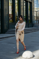 A brunette in a beige dress and glasses in the middle of the city on the background of buildings