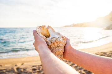 Fototapeta na wymiar Hands of a woman holding a large seashell on a beach at sunset. In the background you can see the sky and the sea in summer.