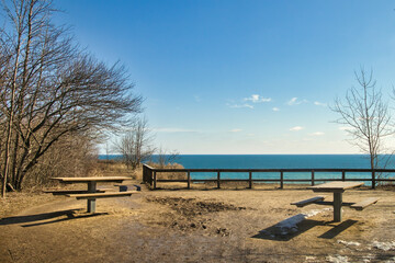 Fototapeta na wymiar Under a sunny blue sky in early Spring, two empty picnic tables face a peaceful Lake Michigan from atop a bluff at Lion's Den Gorge, near Grafton, WI.