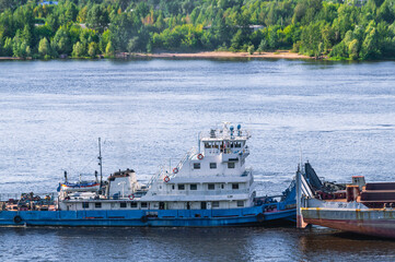 A tugboat pushes a large barge with cargo along a wide river. In summer, the tugboat goes down the...