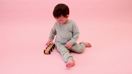 Happy toddler baby is playing with a toy car against a pink background. Child boy rolls a wooden toy car. Kid age one year eight months, full height