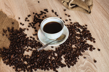 A cup of hot coffee in a plate with a spoon. Coffee beans are scattered on a wooden light brown table.