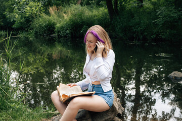 A young schoolgirl with stylishly dyed hair, in denim shorts and a white shirt, sits on a stone near a picturesque river with a book in her hands, laughs and talks on the phone.