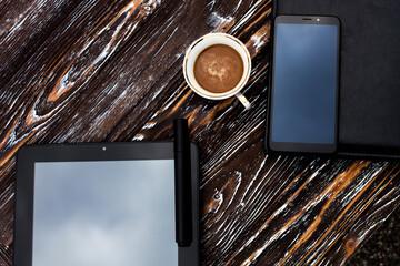 A cup of aromatic coffee, a black tablet, a mobile phone, a notebook with a leather cover and a pen...