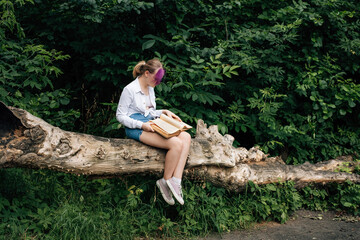 A young schoolgirl with stylishly dyed hair, with a purple streak of hair, in denim shorts and a white shirt, sits on a tree and reads a big book.