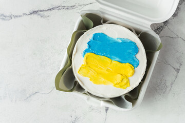 Patriotic cake decorated with yellow and blue Ukrainian flag. Small trendy bento cake in the white gift box. Korean style little cake on the white marble background.