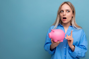 Obraz na płótnie Canvas pensive blond girl holding a piggy bank with a stock thinks where to spend money on a blue isolated background