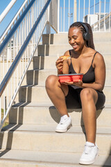 Naklejka premium smiling young black Afro-Latin girl holds healthy food and listens to music with headphones outdoors. african american woman in sportswear sitting on steps 
