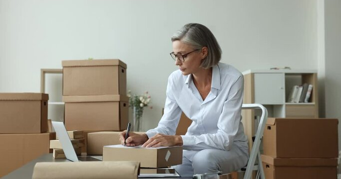 Attractive mature businesswoman, small business owner prepare goods for sending, writes customer data, shipping parcel box using fast and safe transporting services. Shipment, delivery, dropshipping