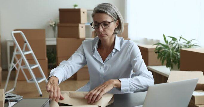 Mature female retail seller, on-line store owner prepares goods for shipment, packing box in wrapping paper, working from storage room. Trade, small business entrepreneurship, fast delivery services