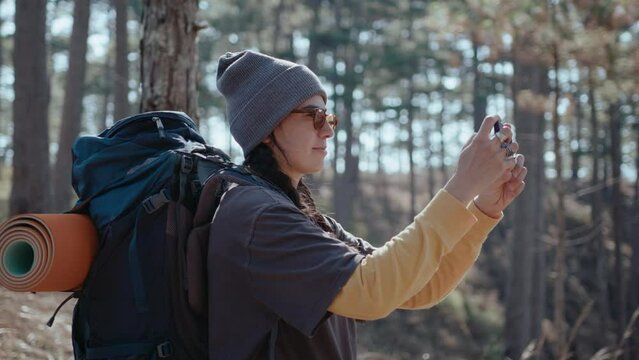Girl with backpack standing in woods, photographing nature with smartphone, hiking on summer day. Medium shot, side view