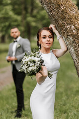 Beautiful young bride standing on lane in summer park, groom with bouquet in background. Beautiful evening white dress