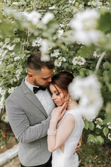 the bride in a white evening dress poses with the groom with a beautiful hairstyle, the groom gently wraps the bride and leans against her against the background of spring flowers.