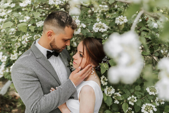 the bride in a white evening dress poses with the groom with a beautiful hairstyle, the groom gently wraps the bride and leans against her against the background of spring flowers.