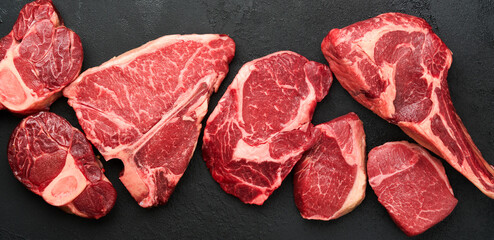 Raw prime steaks. Variety of fresh black angus prime meat steaks T-bone, New York, Ribeye, Striploin, Tomahawk cutting board on black or dark background. Set of various classic steaks. Top view.