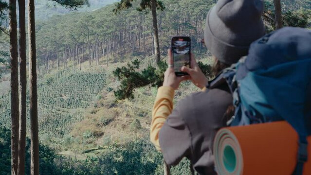 Female hiker with backpack photographing scenic mountain forest with smartphone, standing on the hill, traveling alone in nature. Back shot, copy space