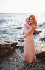 Fototapeta na wymiar Young adult beautiful single mother in a pink dress with long curls hair makeup, holding her baby girl in a pink bodysuit background of the sea in the summer at sunset. Happy Motherhood, mothers Day