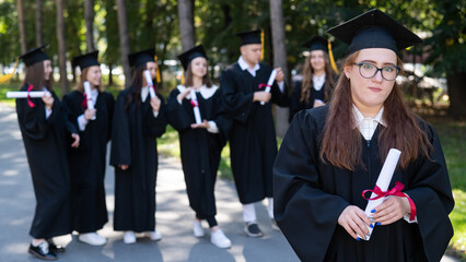 Portrait of a young caucasian woman in glasses and a graduate gown against the background of classmates. A group of graduate students outdoors.