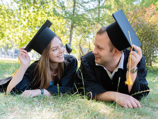 Happy guy and girl look at each other lying on the lawn in a black graduate gown and an academic cap of the university