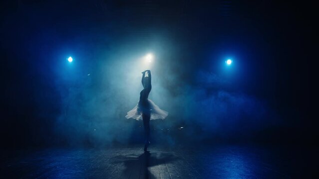 Silhouette of graceful ballerina in blue backlight of spotlights. Young ballet dancer twirling on toes in pointe shoes in stage smoke