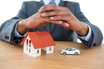 Businessman protecting house model and car with hands on table