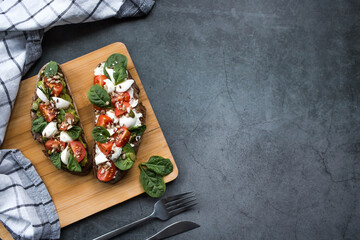 Bruschetta (sandwiches) with cherry tomatoes, mozzarella cheese and herbs on a cutting board on a dark background. Traditional Italian snack.