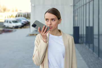 Smiling woman with smartphone in hand recording voice message to colleague outdoors. Smiling female employee in official wear walking near business center