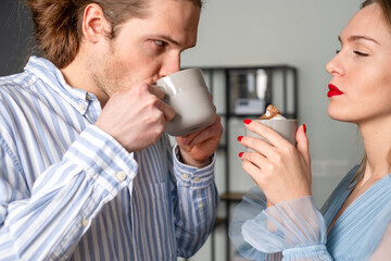 A worried man and a beautiful, proud woman with red lips and nails, both in light blue clothes, are drinking coffee. The bookshelf is in the background.