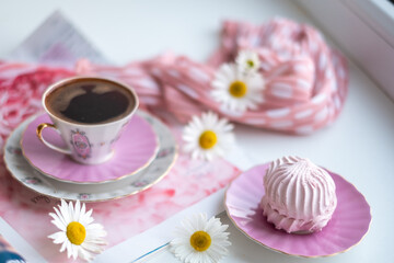 Fototapeta na wymiar Black coffee in a gorgeous fragile coffee cup on a soft pink napkin and puffy airy pink marshmallow in a saucer, a stylish polka dot scarf and beautiful sunny daisy flowers on the white table