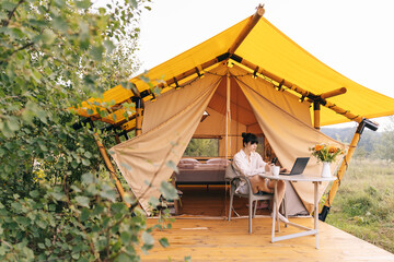 young woman working on a laptop while sitting near a big tent in nature