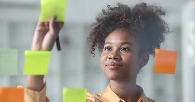 Young African American stylish woman working on project plan using sticky papers notes on glass wall, people meeting to share idea, positive thinking workshop and business design planning concepts.