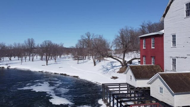 Aerial shot of a swirling river next to an old red and white grain mill in winter