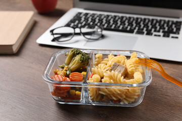Container of tasty food, fork, laptop and glasses on wooden table. Business lunch