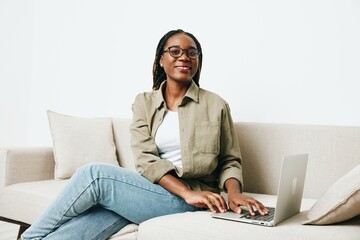 Stylish African American woman business freelancer working sitting on couch at home in a laptop and phone, business calls and correspondence, home clothes and eyeglasses, light interior background.