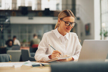 Woman office worker working on laptop and making notes while sitting in coworking