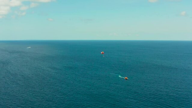 People flying on a colorful parachute towed by a motor boat, aerial view. Parasailing in blue sky.