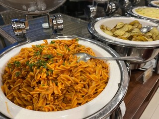 Big portion of rotini or spiral pasta with tomato sauce being served in chafing dish.