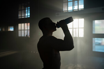 Topless muscular sportsman drinking water from a sports bottle in abandoned warehouse after doing...