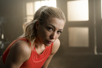 Fototapeta premium Tired athlete leans forward and looks at the camera during a rest break in a gym. Close-up fitness portrait of a woman in a red training top, with warm window light behind her.