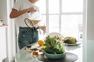 Young slim woman in white t-shirt and blue jeans pouring fruit smoothie healthy food in kitchen at home