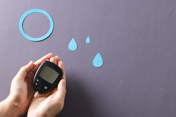 Hands of caucasian woman holding glucometer over blue drops and ring on grey background, copy space