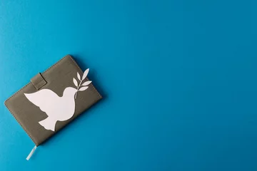 Poster Close up of white dove with leaf over notebook and copy space on blue background © vectorfusionart