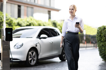 Businesswoman using tablet, walking while recharging her electric vehicle with charging station at...