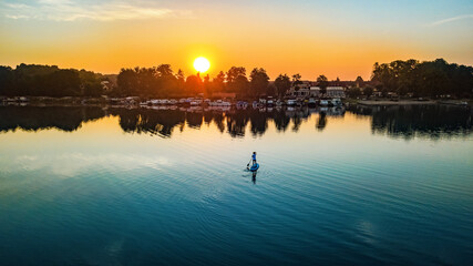 Obraz premium Woman paddling on SUP board on beautiful lake during sunset or sunrise, standing up paddle boarding morning adventure in Germany lake district Mecklenburg 