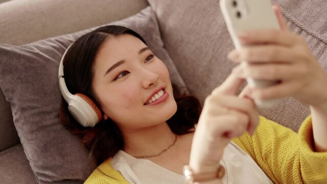 Happy woman, headphones and phone in a living room with social media, music and texting. Home, sofa and smile of a Asian female and gen z student feeling relax from audio and web podcast on couch